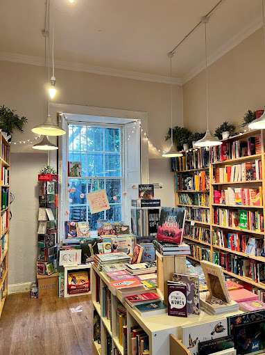 A cosy, well-lit bookshop interior with white walls and wooden flooring. Shelves filled with colourful books line the walls, and a central display table showcasing various book titles. A large window at the back lets in natural light, and fairy lights add