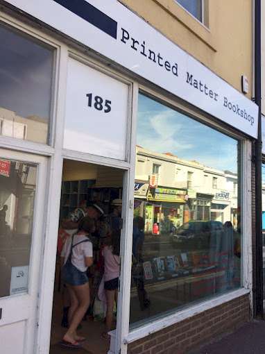 The front of Printed Matter Bookshop, featuring large windows displaying books and a sign with the bookshop's name and address (185). Customers can be seen inside the shop, adding to its inviting atmosphere.