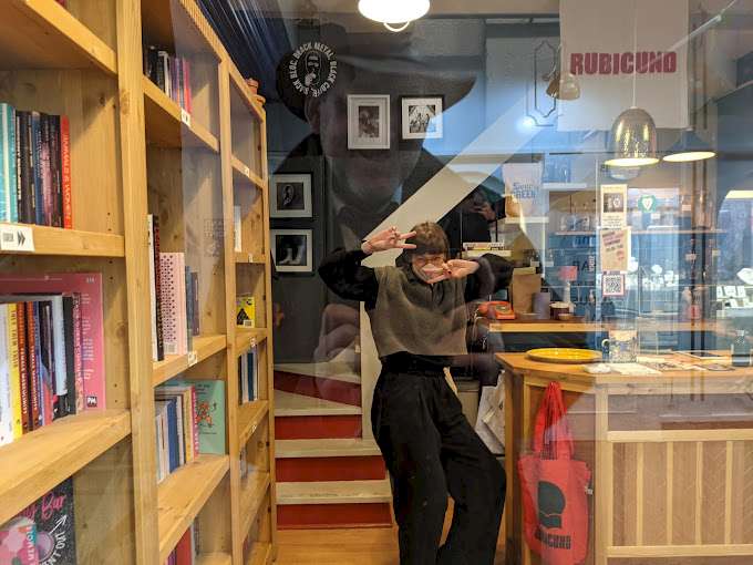 A playful moment captured through the glass of Rubicund Bookshop, showcasing shelves filled with books. A person poses playfully behind the glass of a bookshop, making a peace sign with both hands. Shelves filled with books are visible on the left, and th