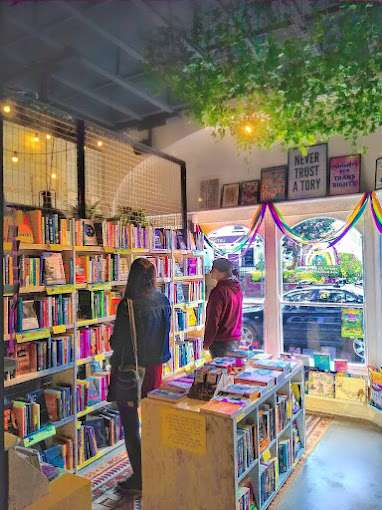 The interior of Shelf Life Books and Zines in Cardiff, showcasing shelves filled with books, colourful decorations, and a cosy atmosphere. Two people browse the collection, and rainbow streamers hang in the window. © Rani