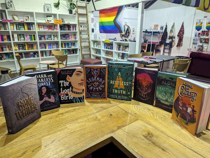 A colourful selection of books displayed on a wooden table inside Paperxclips, an LGBTQIA+ bookshop in Belfast. The background features bookshelves filled with books, a seating area, and a pride flag on the wall. © Paperxclips Books