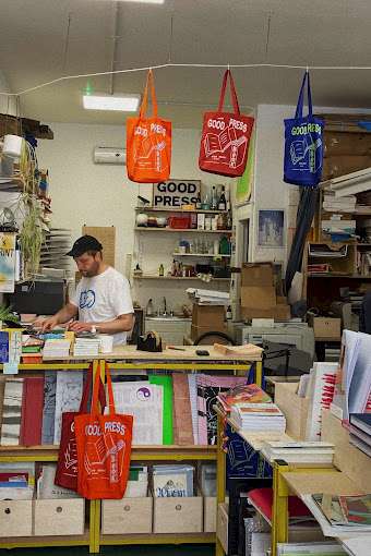 The interior of Good Press in Glasgow, featuring shelves filled with books and zines, and colourful tote bags hanging above. A staff member is seen working behind the counter. © Iris Fu