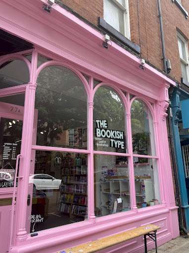 The front of The Bookish Type bookshop in Leeds, featuring a bright pink facade with large windows displaying books. The shop sign reads "The Bookish Type," and a bench is located outside.