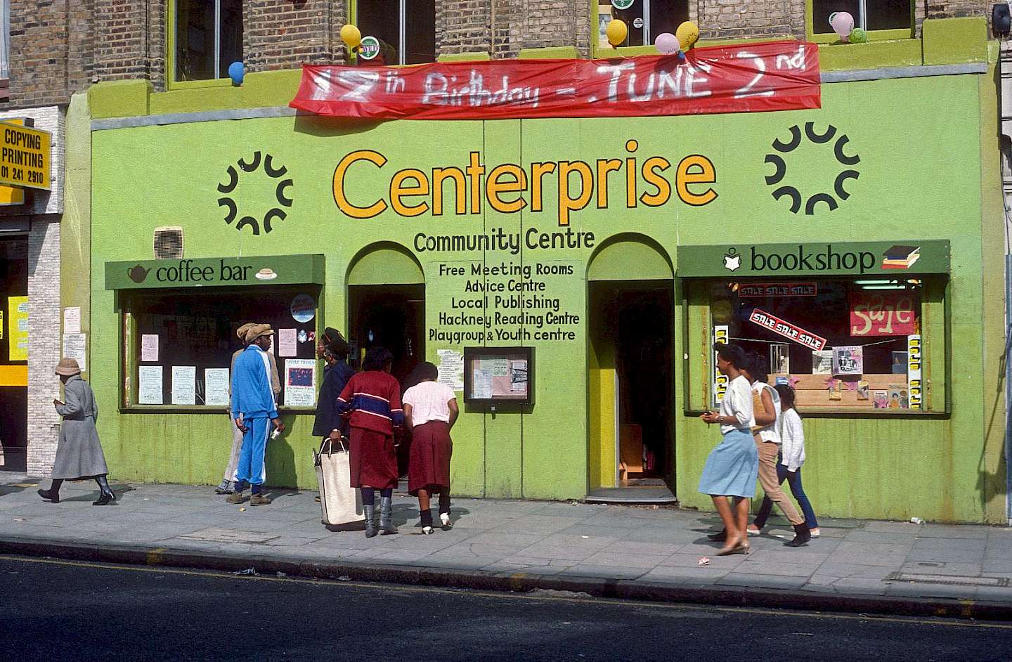 A photo of the exterior of Centerprise Community Centre in Hackney, London, from the 1980s. The building is painted green and features a coffee bar and bookshop. People are walking by and interacting in front of the entrance, and a banner celebrating an a