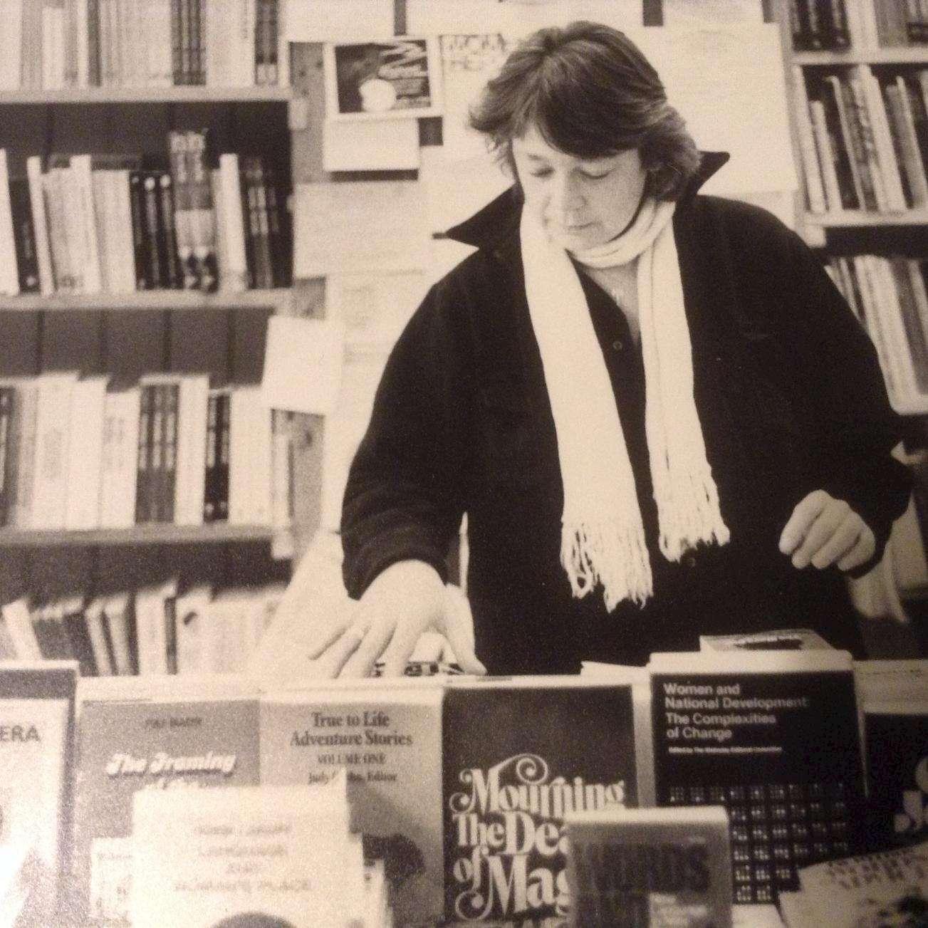 A black-and-white photo of a woman with short hair and a scarf, standing behind a display of books in a bookstore. The shelves behind her are filled with books and notices.