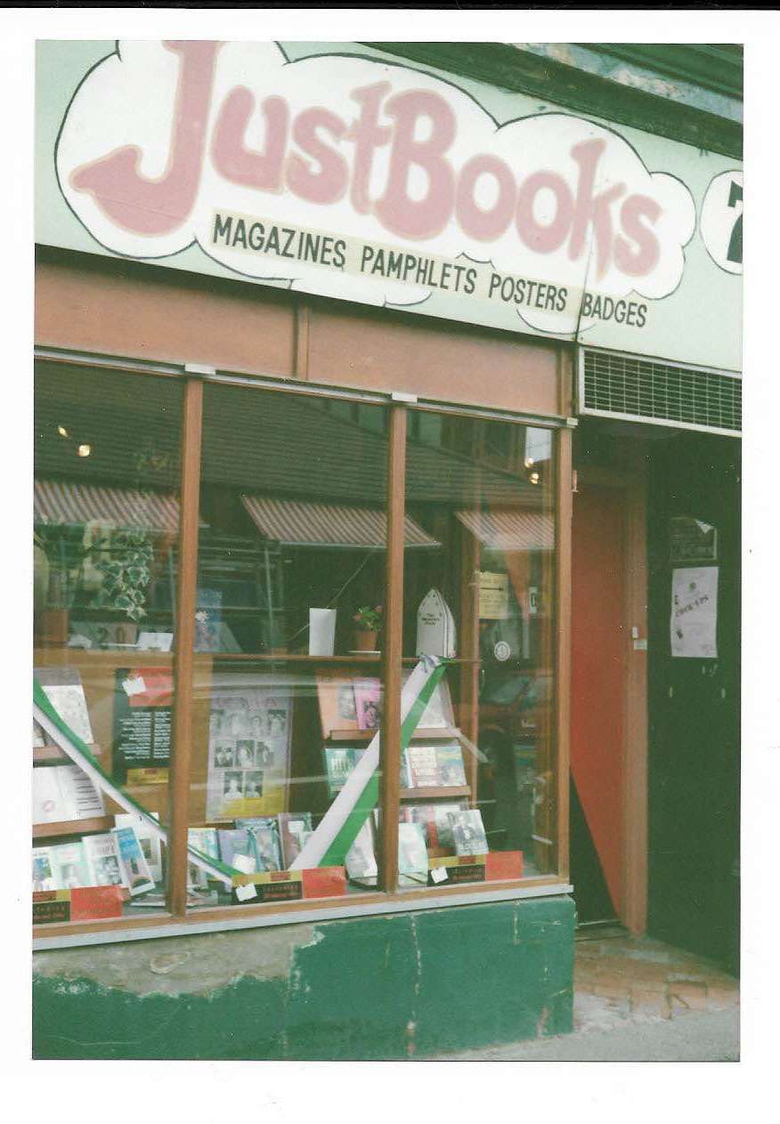 A photograph of the Just Books storefront in Belfast, showcasing large windows filled with books, magazines, pamphlets, and posters. The sign above the shop reads "Just Books" with additional text listing "Magazines, Pamphlets, Posters, Badges."