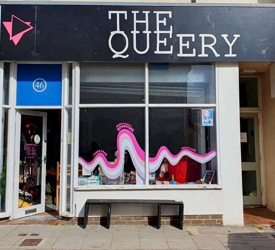 The front of The Queery in Brighton, featuring a bold black sign with the shop's name in white letters. The window displays a wave that reads "Queer Radical Bookshop" and "Vegan Café." The entrance is to the left, showing part of the interior.