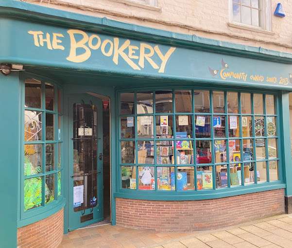 The front of The Bookery in Crediton, Devon, featuring a large, curved window display filled with books. The shopfront is painted green with the bookshop's name in yellow letters. © The Bookery