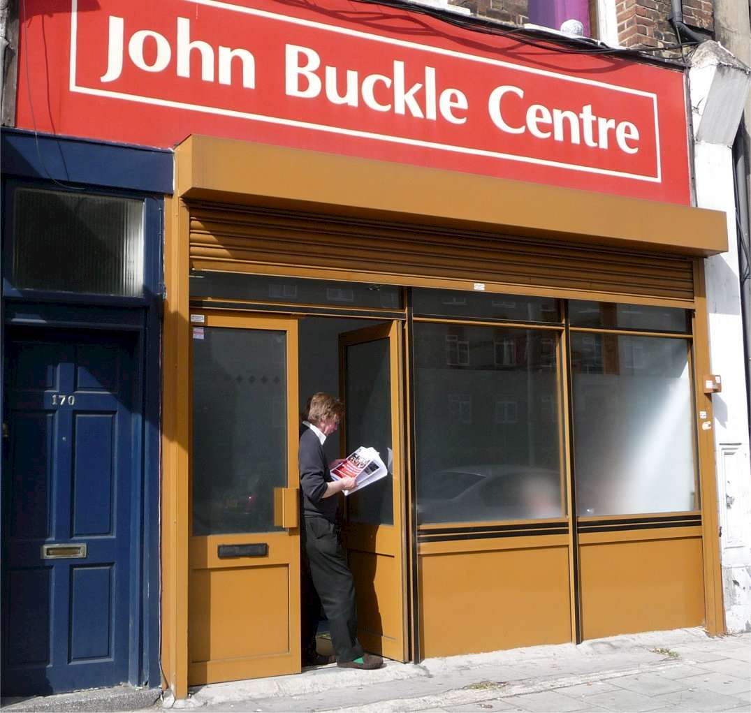 A man standing outside the John Buckle Centre, reading a newspaper. The building has a red sign with white text and brown doors and windows. © John Buckle Centre